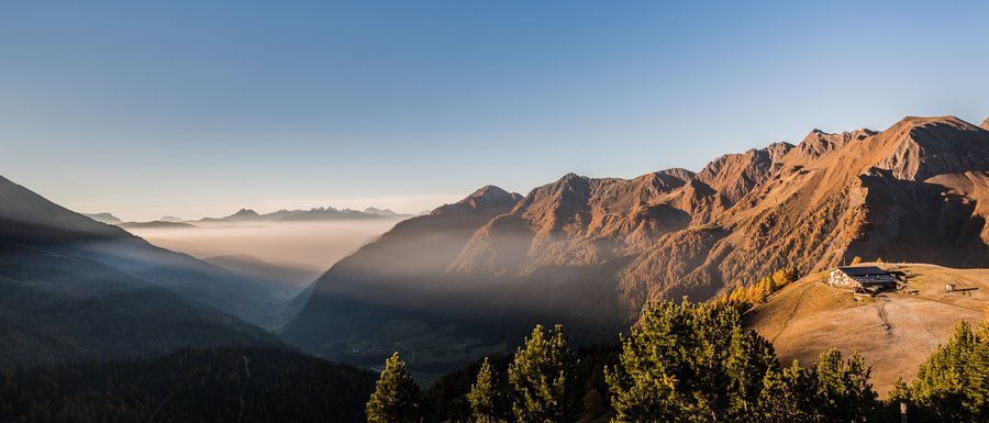 Benvenuti nell’Area Vacanze Sci e Malghe Rio Pusteria! L'immagine mostra un pittoresco paesaggio montano all'alba o al tramonto, con valli profonde coperte di nebbia e pendii di montagna illuminati dal sole che brillano in calde tonalità arancioni. Su una cresta montuosa a destra dell'immagine si trova un rifugio di montagna, circondato da colline erbose e alberi coniferi.