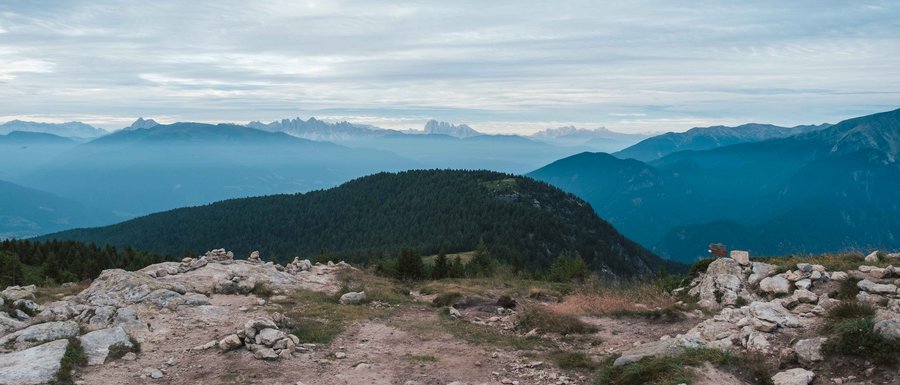 Benvenuti nell’Area Vacanze Sci e Malghe Rio Pusteria! L'immagine mostra un paesaggio montano con più livelli di colline e montagne che si estendono in lontananza. In primo piano c'è una superficie rocciosa, mentre sullo sfondo si vedono colline boschive e catene montuose più lontane sotto un cielo nuvoloso.