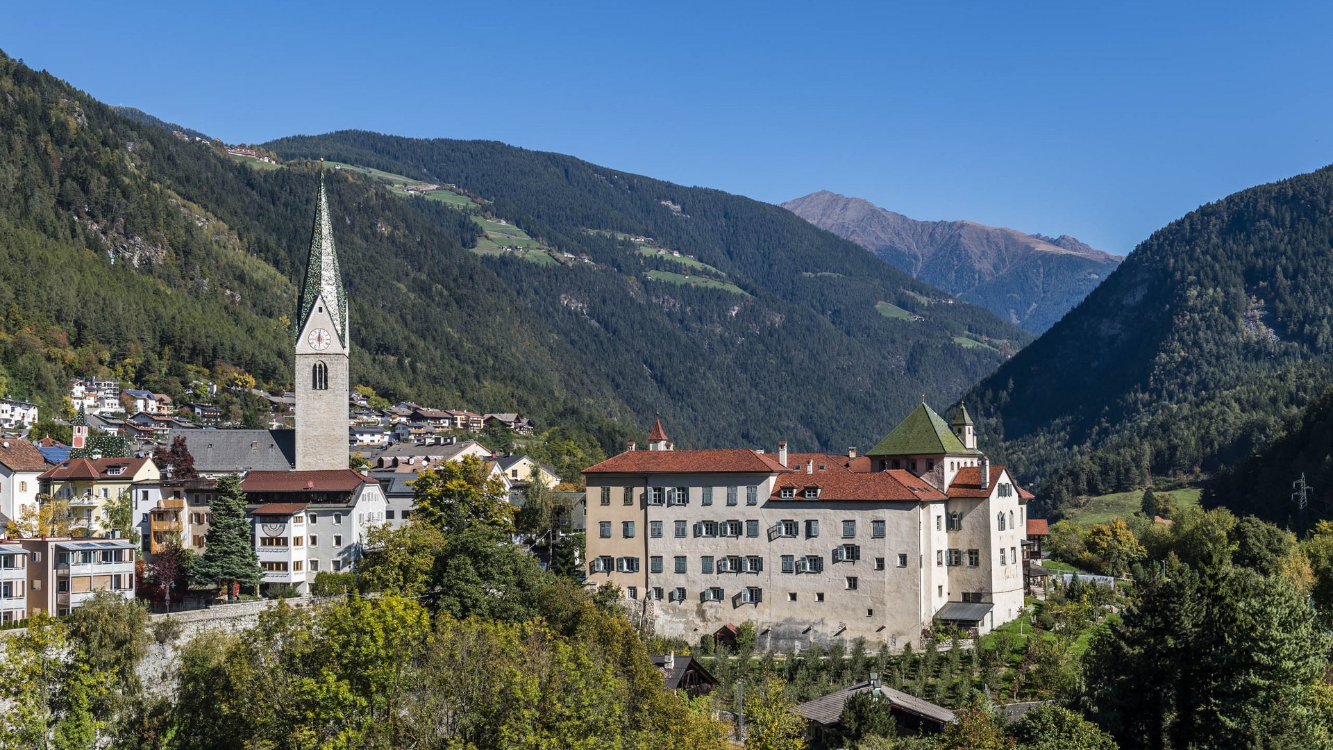 Alla scoperta di Rio di Pusteria L'immagine mostra una pittoresca cittadina in una valle, circondata da colline e montagne boscose. Al centro si trova una chiesa con una torre alta e appuntita, e accanto c'è un grande edificio con tetti rossi e molte finestre.