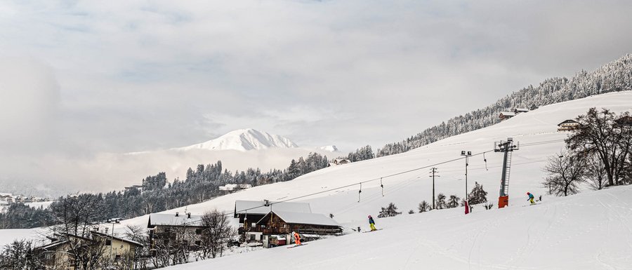 Benvenuti nell’Area Vacanze Sci e Malghe Rio Pusteria! L'immagine mostra un paesaggio innevato con una seggiovia e diversi sciatori su una pista. Sullo sfondo si vedono colline e montagne innevate, parzialmente coperte da nuvole.