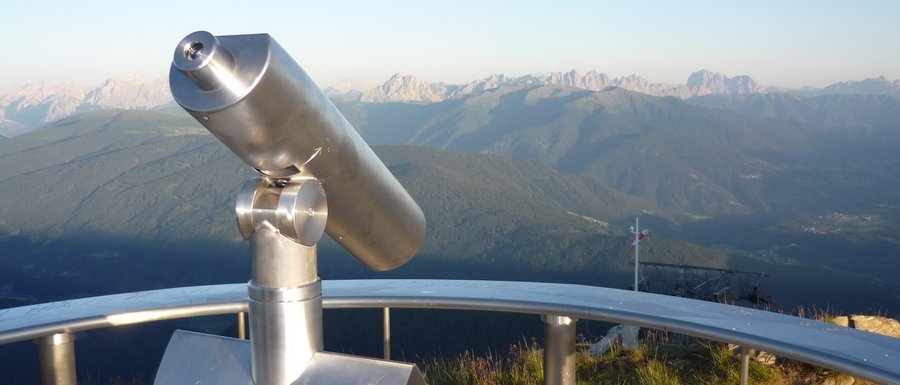 Benvenuti nell’Area Vacanze Sci e Malghe Rio Pusteria! L'immagine mostra un binocolo fissato permanentemente su un punto panoramico, rivolto verso un paesaggio montano ampio. Sullo sfondo si vedono catene montuose e valli sotto un cielo azzurro limpido.