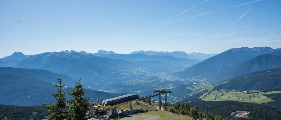 Benvenuti nell’Area Vacanze Sci e Malghe Rio Pusteria! L'immagine mostra un'ampia paesaggio montano sotto un cielo azzurro e limpido. In primo piano c'è una stazione della funivia sulla vetta di una montagna, mentre sullo sfondo si vedono diverse catene montuose e una valle con villaggi sparsi e boschi. Due parapendii volano in alto nel cielo sopra la scena.
