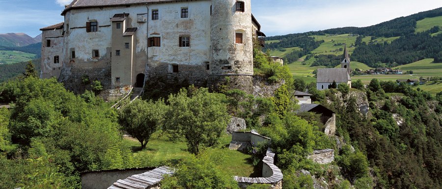 Benvenuti nell’Area Vacanze Sci e Malghe Rio Pusteria! L'immagine mostra un vecchio castello su una collina verde, circondato da alberi e da un muro di pietra. Sullo sfondo si vede una chiesa e un paesaggio rurale.