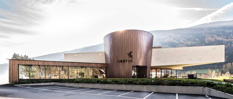 Benvenuti nell’Area Vacanze Sci e Malghe Rio Pusteria! L'immagine mostra un edificio moderno con una facciata rotonda color rame e grandi finestre di vetro. Sull'edificio è visibile la scritta "Capriz" e davanti c'è un parcheggio vuoto, mentre sullo sfondo si riconoscono colline e montagne boschive.
