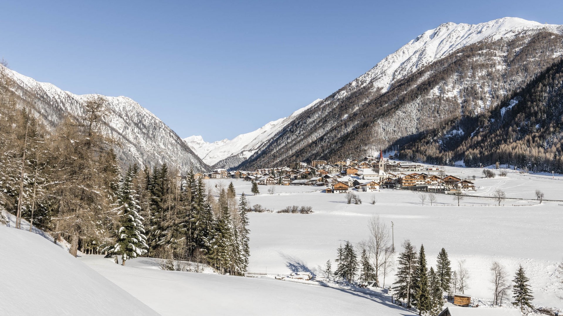 Valles & il villaggio alpino più bello dell’Alto Adige L'immagine mostra un paesaggio invernale con un villaggio in una valle, circondato da montagne innevate. In primo piano si vedono alberi e colline coperte di neve, mentre il cielo è limpido e blu.