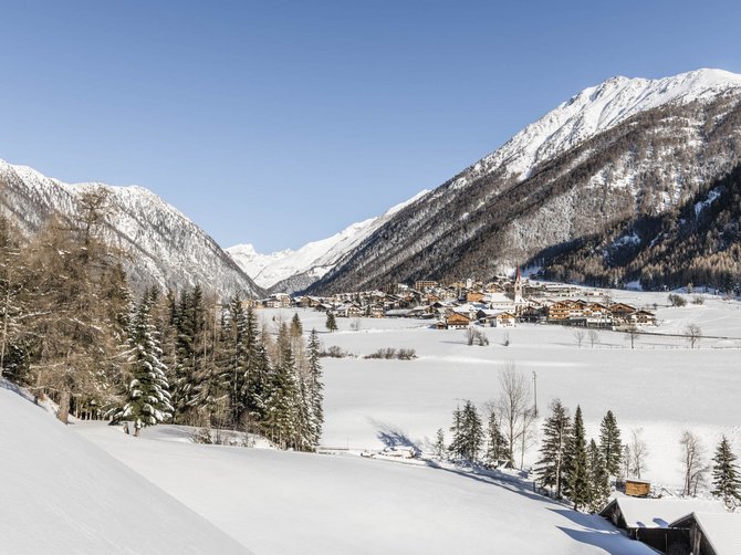 Il paradiso escursionistico di Rodengo L'immagine mostra un paesaggio invernale con un villaggio in una valle, circondato da montagne innevate. In primo piano si vedono alberi e colline coperte di neve, mentre il cielo è limpido e blu.