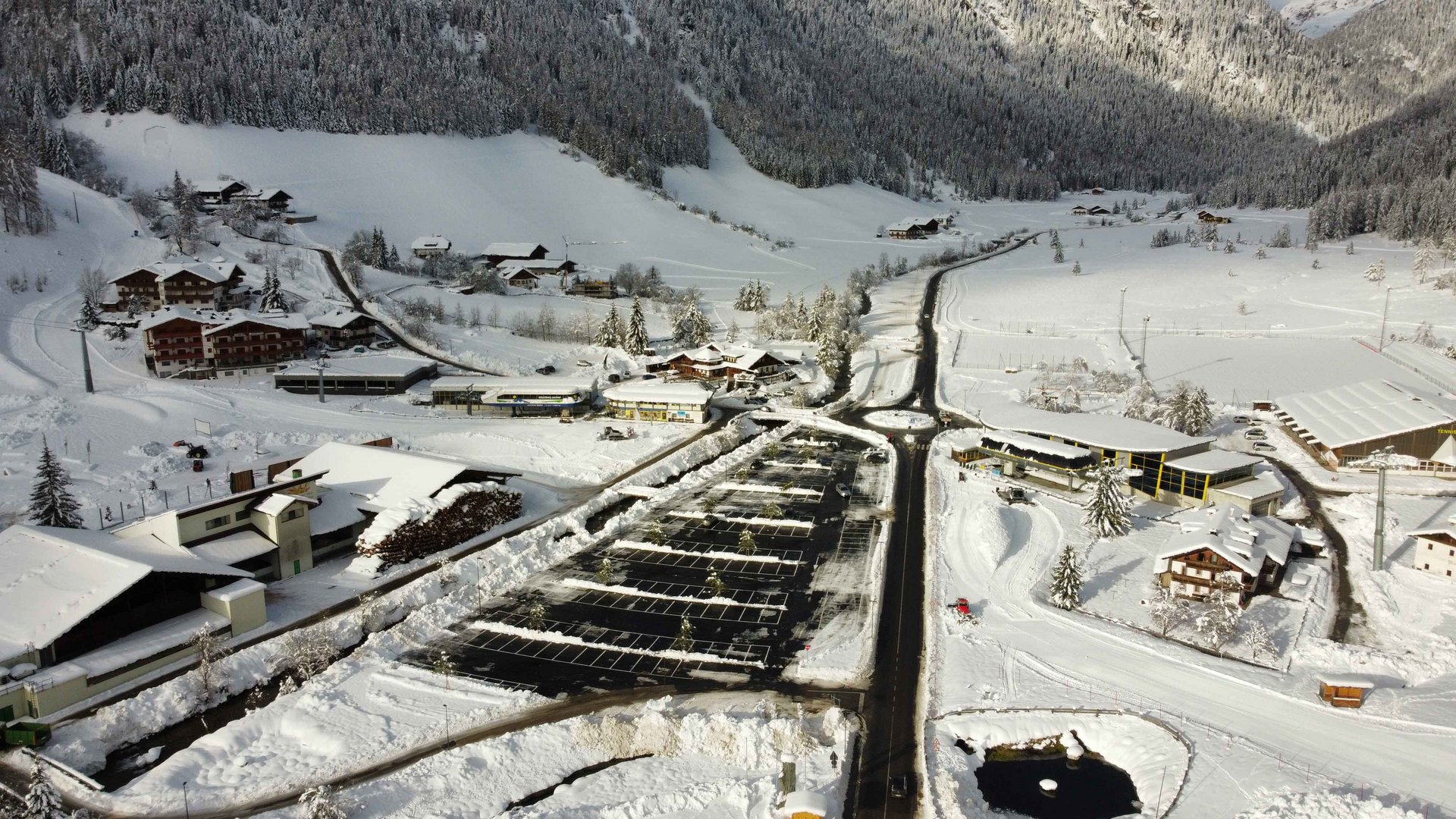 Valles & il villaggio alpino più bello dell’Alto Adige L'immagine mostra un villaggio innevato visto dall'alto. Le strade e le case sono coperte di neve, e in primo piano si vede un parcheggio con posti auto sgomberati. Sullo sfondo si estende un pendio montano boscoso.