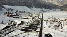 Valles & il villaggio alpino più bello dell’Alto Adige L'immagine mostra un villaggio innevato visto dall'alto. Le strade e le case sono coperte di neve, e in primo piano si vede un parcheggio con posti auto sgomberati. Sullo sfondo si estende un pendio montano boscoso.