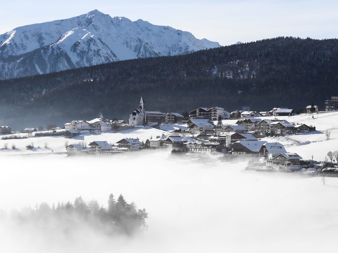 Il paradiso escursionistico di Rodengo L'immagine mostra un villaggio innevato in una valle, circondato da una fitta nebbia. Sullo sfondo si vedono alte montagne innevate e un pendio boscoso. Una chiesa spicca al centro del villaggio e il cielo è sereno e azzurro.