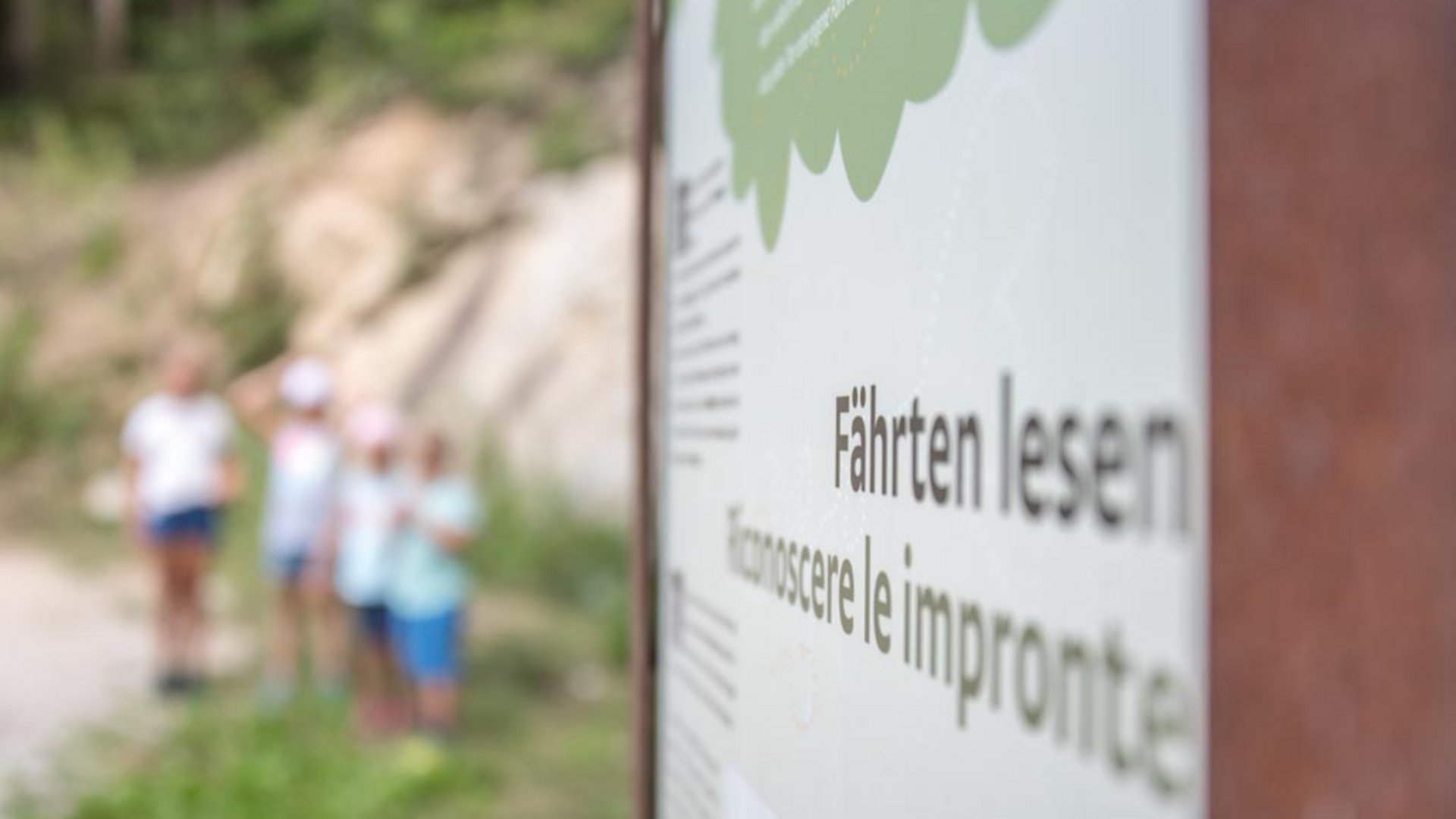 Il Sentiero degli Elfi a Vandoies di Sopra L'immagine mostra un cartello con la scritta "Fährten lesen" e la traduzione italiana "Conoscere le impronte". Sullo sfondo si intravedono sfocati alcuni bambini in una zona boschiva.