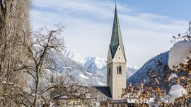 Alla scoperta di Rio di Pusteria L'immagine mostra una chiesa con un alto campanile verde in un villaggio innevato. Sullo sfondo si vedono montagne coperte di neve e un cielo azzurro, mentre in primo piano ci sono alberi spogli e cespugli coperti di neve.