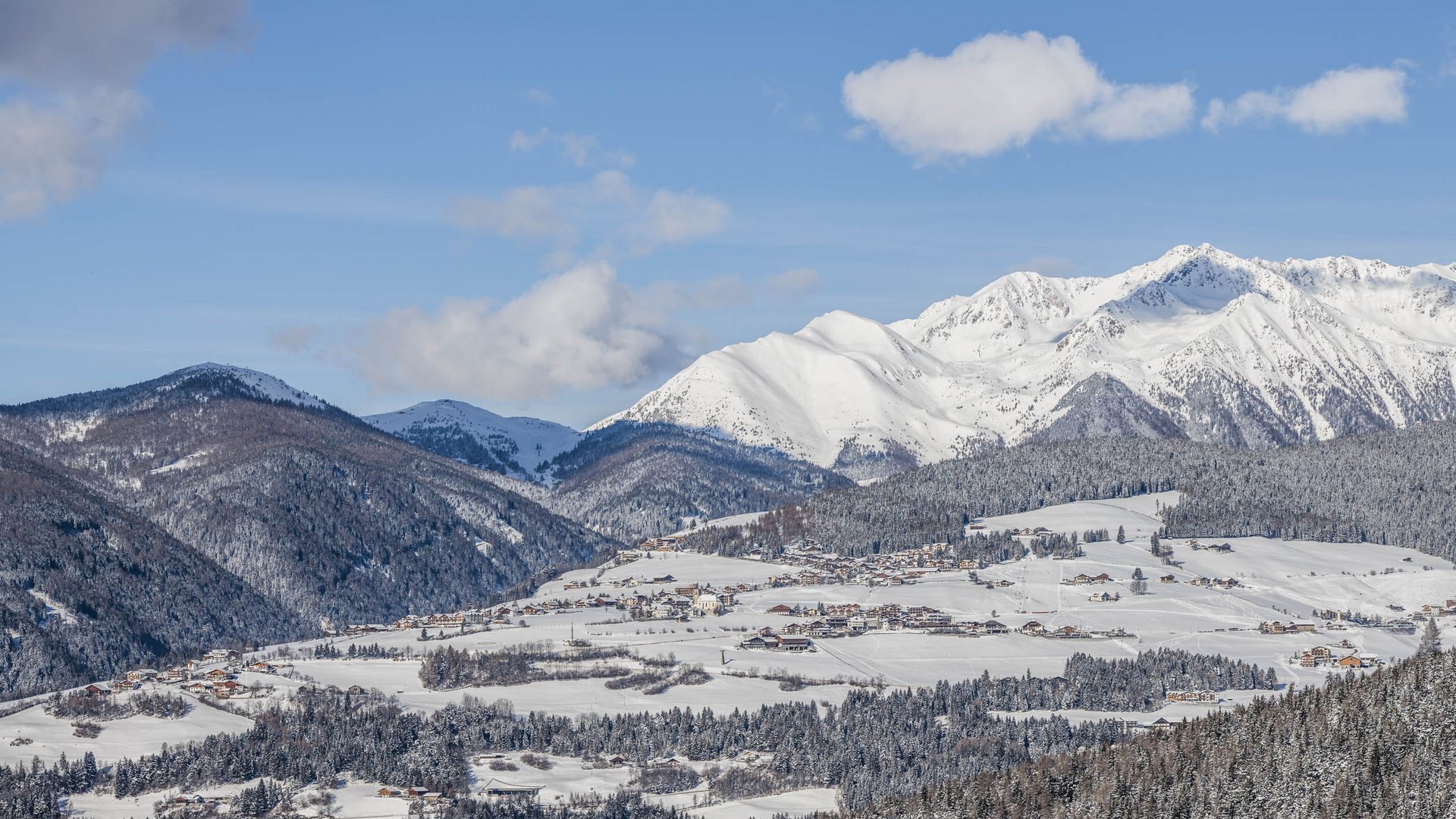 Conoscete già Maranza? L'immagine mostra un paesaggio montano innevato con un villaggio in una valle. Le montagne circostanti sono anch'esse coperte di neve e si ergono maestosamente sotto un cielo limpido e azzurro. Il villaggio e i pendii boscosi sono avvolti da una neve fresca, e si possono vedere isolate costruzioni e strade.