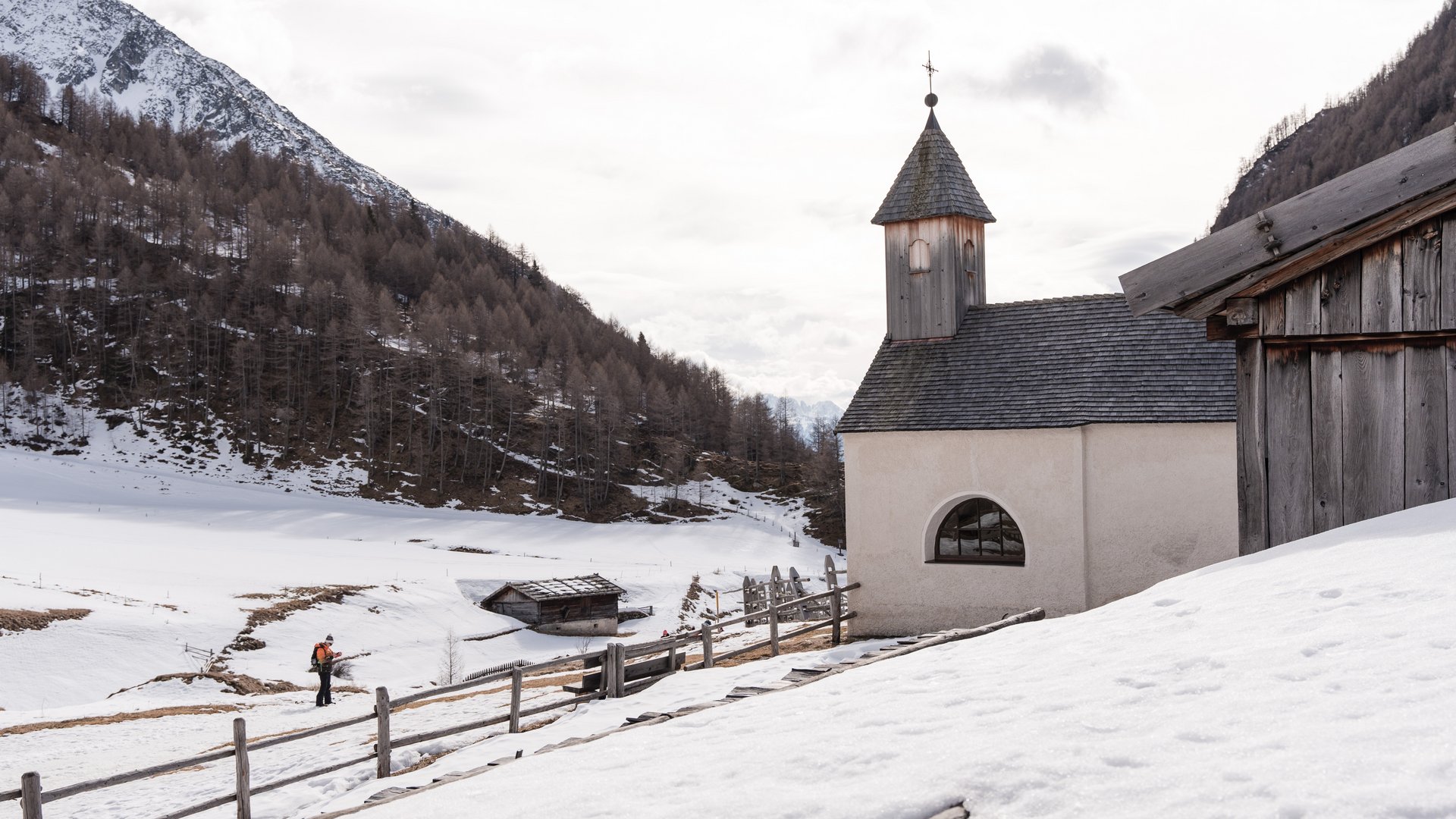 Malga Fane, il più bel villaggio alpino dell’Alto Adige L'immagine mostra un paesaggio innevato con una piccola cappella semplice, che ha un tetto di scandole di legno e una torre con una croce. Sullo sfondo si vedono colline e alberi coperti di neve, mentre un escursionista cammina su un sentiero lungo una recinzione di legno.
