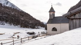 Malga Fane, il più bel villaggio alpino dell’Alto Adige L'immagine mostra un paesaggio innevato con una piccola cappella semplice, che ha un tetto di scandole di legno e una torre con una croce. Sullo sfondo si vedono colline e alberi coperti di neve, mentre un escursionista cammina su un sentiero lungo una recinzione di legno.