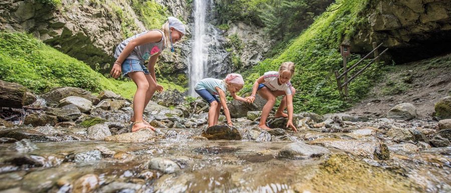 Benvenuti nell’Area Vacanze Sci e Malghe Rio Pusteria! L'immagine mostra tre bambini che giocano a piedi nudi in un ruscello poco profondo. Sullo sfondo si trova una grande cascata che scende da una parete rocciosa ripida, circondata da una rigogliosa foresta verde.