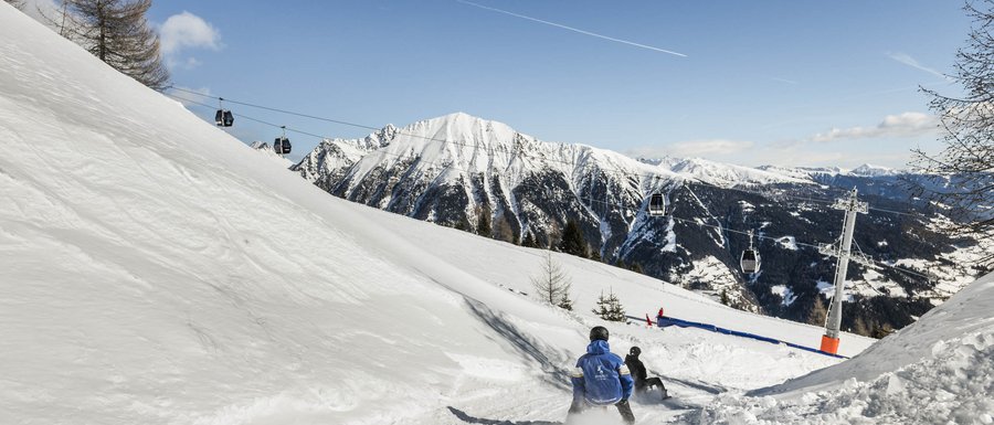 Benvenuti nell’Area Vacanze Sci e Malghe Rio Pusteria! Persone che slittano su pista innevata con funivia e montagne sullo sfondo