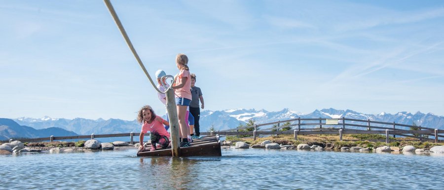 Benvenuti nell’Area Vacanze Sci e Malghe Rio Pusteria! L'immagine mostra diversi bambini che stanno su una piccola zattera, tirata con una corda su uno stagno poco profondo. Sullo sfondo si vedono delle montagne che completano il cielo azzurro limpido e il paesaggio pittoresco.