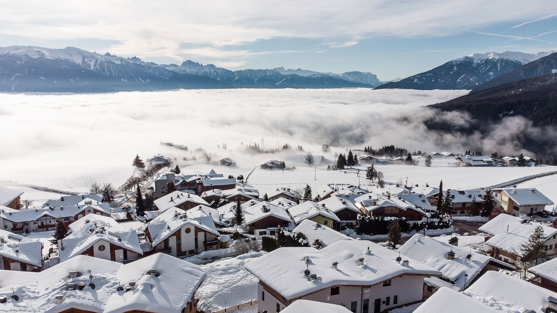 Conoscete già Maranza? L'immagine mostra un villaggio con case innevate su una collina, circondato da una copertura di nebbia. Sullo sfondo si vedono imponenti cime montuose innevate sotto un cielo parzialmente nuvoloso.