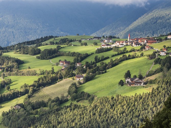 Il paradiso escursionistico di Rodengo L'immagine mostra un paesaggio pittoresco con dolci colline verdi, su cui sono sparse piccole case e fattorie. Sullo sfondo si erge un pendio montano boscoso, mentre nel villaggio si vede una piccola chiesa con il tetto rosso.