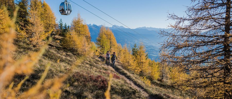 Benvenuti nell’Area Vacanze Sci e Malghe Rio Pusteria! L'immagine mostra una funivia che attraversa un sentiero utilizzato dagli escursionisti. Il paesaggio è autunnale, con alberi dorati, e sullo sfondo si vedono montagne sotto un cielo azzurro limpido.