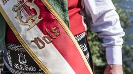 Valles & il villaggio alpino più bello dell’Alto Adige L'immagine mostra un dettaglio di un costume tradizionale. Si vede una fascia rosso-bianco-oro con la scritta "Vals" e un emblema ricamato di un'arpa dorata. Lo sfondo è sfocato e l'attenzione è concentrata sull'abbigliamento e sulla fascia.