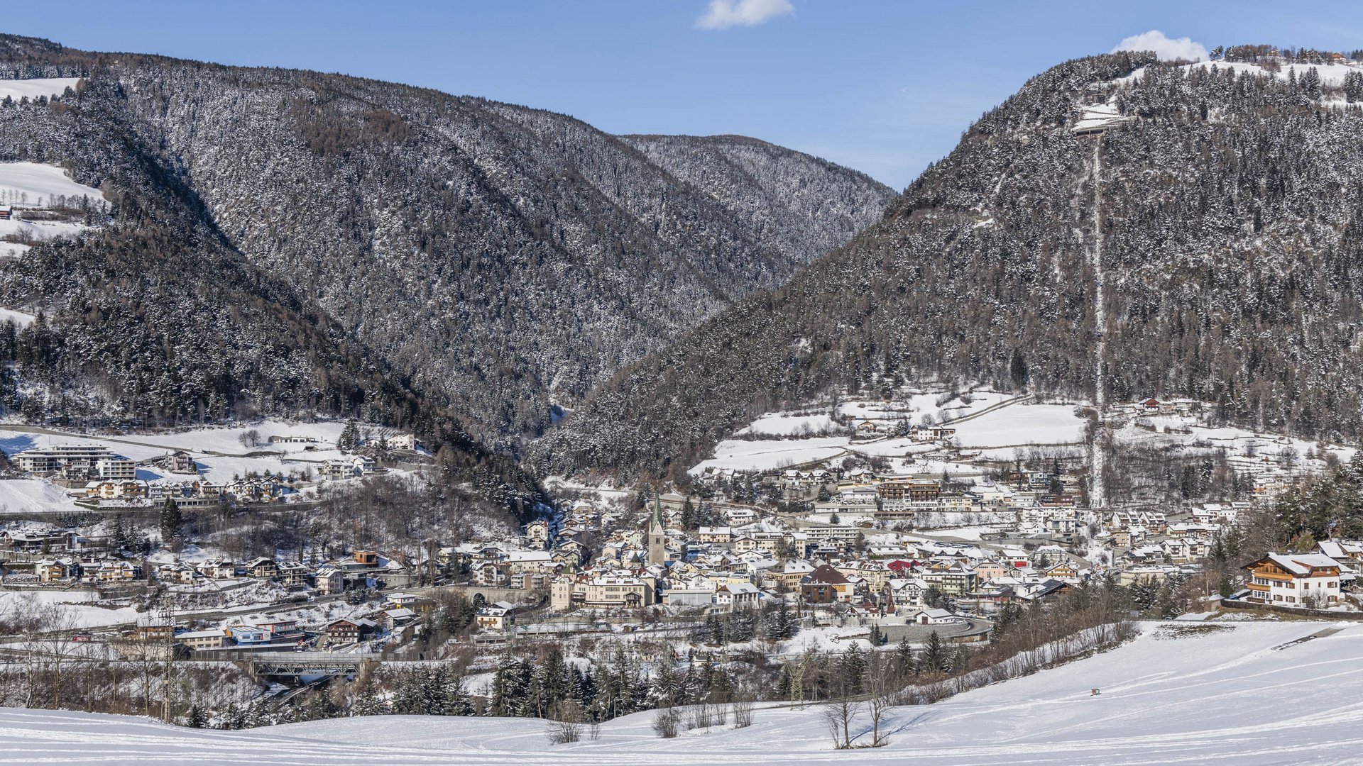 Alla scoperta di Rio di Pusteria L'immagine mostra una città situata in una valle innevata, circondata da colline densamente boscose. Gli edifici sono coperti di neve e sullo sfondo si vedono colline innevate e un cielo azzurro e limpido.
