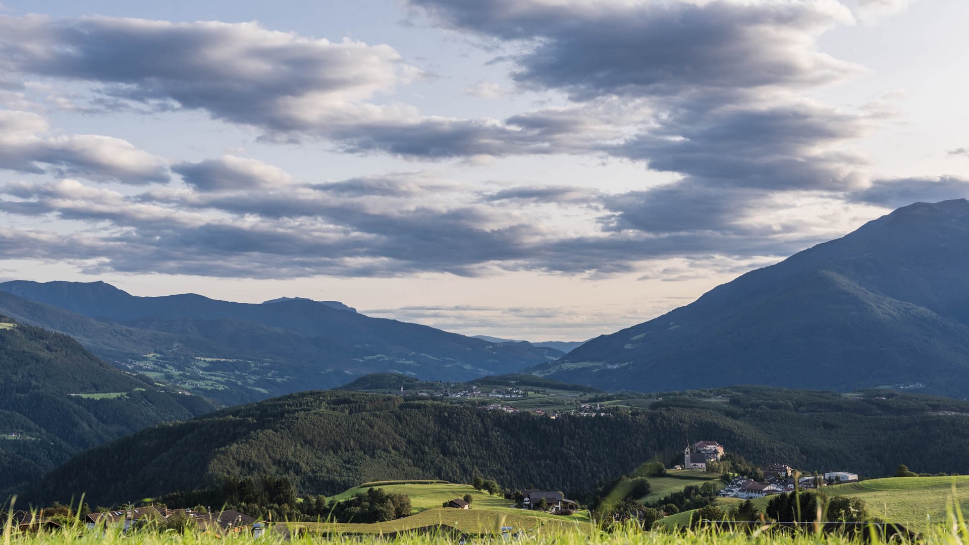 Il paradiso escursionistico di Rodengo L'immagine mostra un vasto paesaggio con dolci colline boschive, sormontate da un cielo leggermente nuvoloso. In primo piano si vedono prati verdi, mentre in lontananza un villaggio si adagia sulle colline.