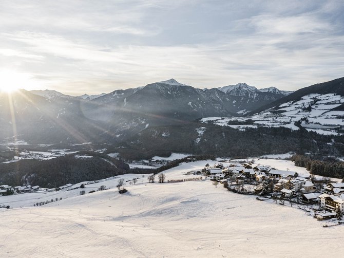 Il paradiso escursionistico di Rodengo L'immagine mostra un paesaggio innevato nelle montagne, poco dopo l'alba. Il sole sorge dietro le montagne e illumina la valle, caratterizzata da campi e colline ricoperte di neve. Sul lato destro dell'immagine si trova un piccolo villaggio, anch'esso coperto di neve. Le montagne sullo sfondo sono in parte boscose e visibili in lontananza.