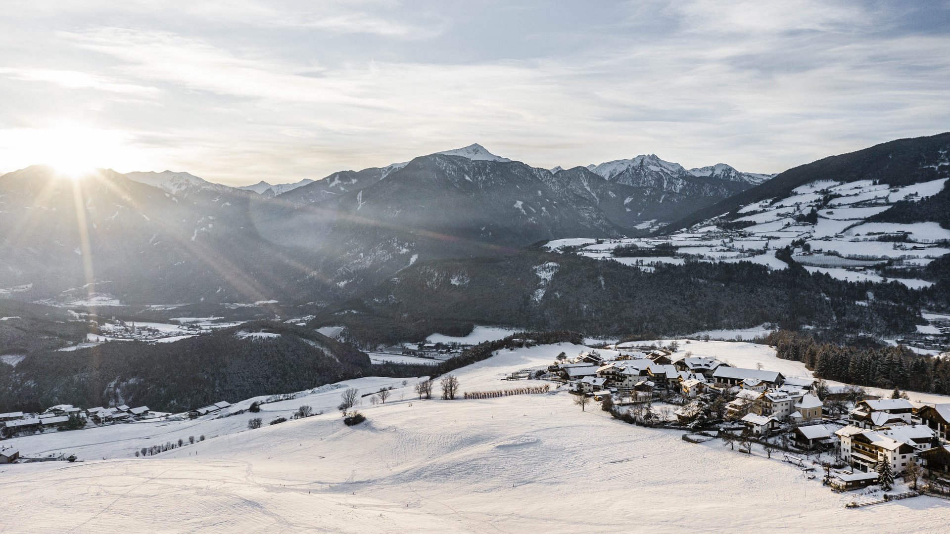 Il paradiso escursionistico di Rodengo L'immagine mostra un paesaggio innevato nelle montagne, poco dopo l'alba. Il sole sorge dietro le montagne e illumina la valle, caratterizzata da campi e colline ricoperte di neve. Sul lato destro dell'immagine si trova un piccolo villaggio, anch'esso coperto di neve. Le montagne sullo sfondo sono in parte boscose e visibili in lontananza.