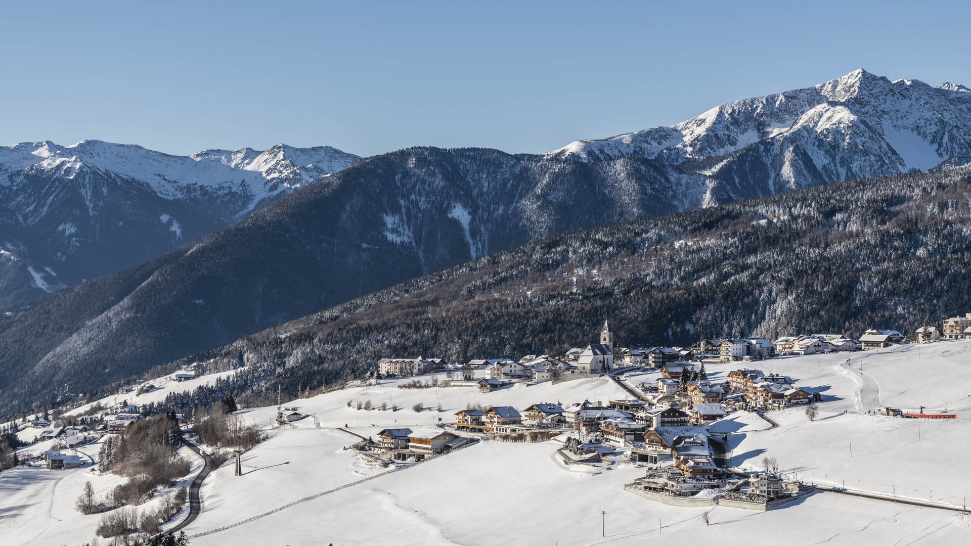 Conoscete già Maranza? L'immagine mostra un villaggio innevato in una valle, circondato da pendii montuosi boscosi e cime innevate. Una chiesa con una torre è posizionata al centro del villaggio, e una strada si snoda attraverso il paesaggio bianco. Il cielo è limpido e blu, creando un'atmosfera invernale soleggiata.