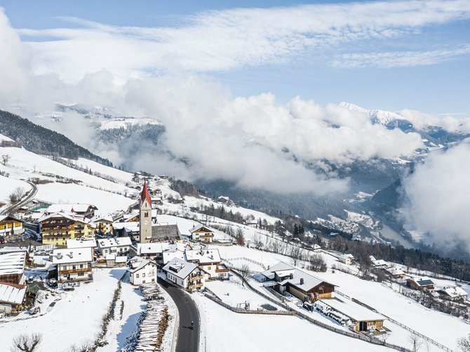Il paradiso escursionistico di Rodengo L'immagine mostra un villaggio di montagna innevato con una chiesa, la cui torre rossa spicca tra le case coperte di neve. Sullo sfondo si vedono colline boschive e nuvole basse che coprono parzialmente la valle sottostante.