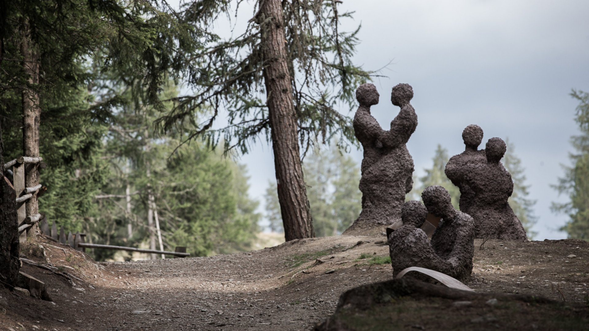 Il Sentiero della Creazione a Rodengo L'immagine mostra diverse sculture astratte di coppie, formate da materiale grezzo, che si trovano in un'area boschiva su un sentiero. Le figure stanno in piedi o sedute vicine l'una all'altra, come se si abbracciassero o fossero in stretto contatto.