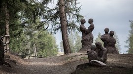 Il Sentiero della Creazione a Rodengo L'immagine mostra diverse sculture astratte di coppie, formate da materiale grezzo, che si trovano in un'area boschiva su un sentiero. Le figure stanno in piedi o sedute vicine l'una all'altra, come se si abbracciassero o fossero in stretto contatto.