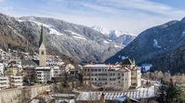 Alla scoperta di Rio di Pusteria L'immagine mostra una città innevata in una valle, circondata da montagne boscose. In primo piano si vede una chiesa con una torre alta e appuntita e diversi edifici storici, mentre sullo sfondo montagne innevate dominano la scena.