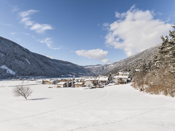 Il paradiso escursionistico di Rodengo L'immagine mostra un paesaggio innevato con un piccolo villaggio nella valle, circondato da montagne coperte di neve. Il cielo è limpido e blu, attraversato solo da qualche nuvola, e in primo piano c'è un albero spoglio sul campo innevato.