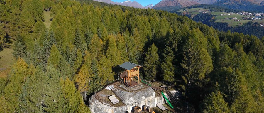 Benvenuti nell’Area Vacanze Sci e Malghe Rio Pusteria! L'immagine mostra una piattaforma panoramica in legno situata su un promontorio roccioso nel mezzo di una fitta foresta. Sullo sfondo si vedono montagne verdi e un cielo azzurro limpido, che offrono una vista ampia sul paesaggio.