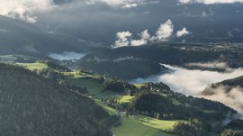 Il paradiso escursionistico di Rodengo L'immagine mostra un paesaggio collinare verde con foreste e prati, attraversato da strade tortuose. La nebbia si insinua nelle valli e avvolge i villaggi e le chiese circostanti, mentre le colline in primo piano sono limpide e soleggiate.