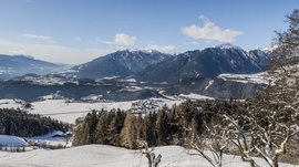 Il paradiso escursionistico di Rodengo L'immagine mostra un ampio paesaggio innevato con dolci colline e villaggi sparsi situati in una valle. Sullo sfondo si ergono maestose montagne, le cui cime sono anch'esse coperte di neve, sotto un cielo azzurro e limpido.