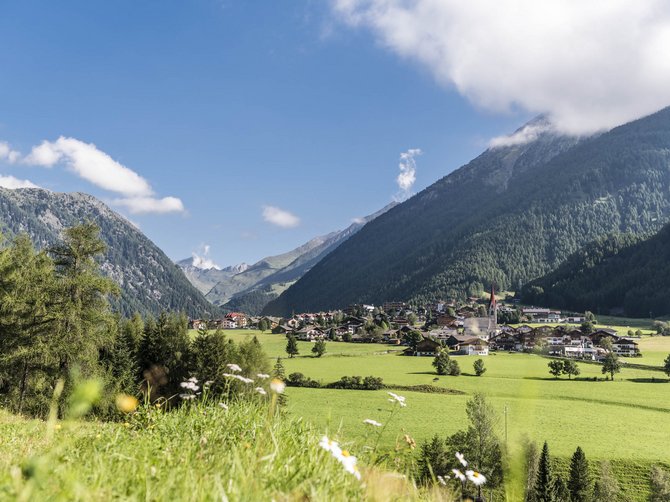Il paradiso escursionistico di Rodengo L'immagine mostra un'idilliaca paesaggio estivo con prati verdi e un villaggio nella valle, circondato da alte montagne boschive. Il cielo è azzurro con alcune nuvole e in primo piano fioriscono sparsi fiori bianchi.