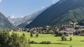 Valles & il villaggio alpino più bello dell’Alto Adige L'immagine mostra un pittoresco villaggio di montagna con case che si adagiano in una valle verde. Sullo sfondo si ergono pendii montani coperti di boschi, mentre una chiesa con una torre rossa caratterizza il paesaggio.