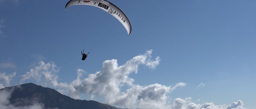Benvenuti nell’Area Vacanze Sci e Malghe Rio Pusteria! L'immagine mostra un parapendio che sorvola in alto un paesaggio montano. Il cielo è limpido e azzurro, con alcune nuvole sparse, e si vede la silhouette di una montagna sullo sfondo.