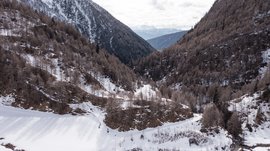 Malga Fane, il più bel villaggio alpino dell’Alto Adige L'immagine mostra una valle innevata circondata da pendii montani coperti di boschi. Gli alberi sono spogli e sullo sfondo si estendono cime innevate visibili in lontananza.
