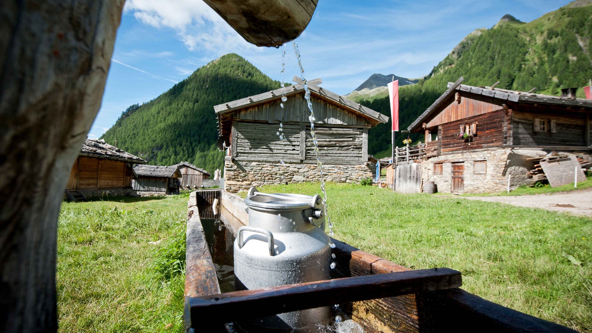 Malga Fane, il più bel villaggio alpino dell’Alto Adige L'immagine mostra un tradizionale pozzo in legno da cui l'acqua scorre in una tinozza. In primo piano c'è una lattiera d'argento nell'acqua, mentre sullo sfondo si vedono diverse capanne rustiche di legno davanti a un paesaggio montano verde.