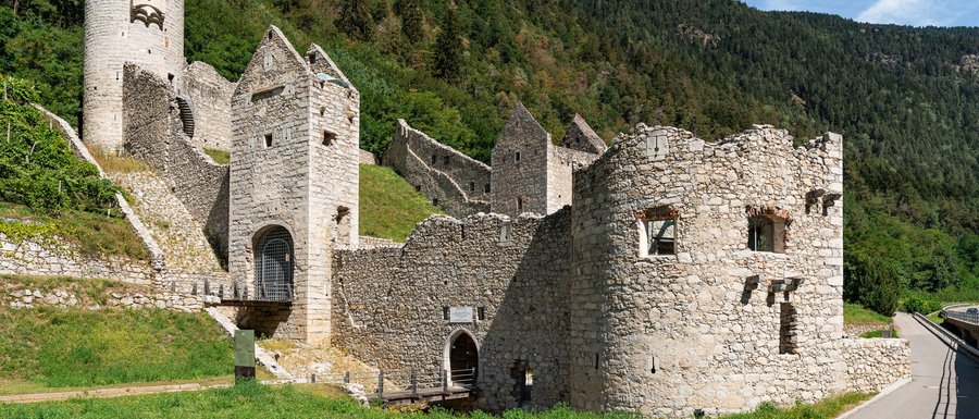 Benvenuti nell’Area Vacanze Sci e Malghe Rio Pusteria! L'immagine mostra un'antica rovina di un castello in pietra con diverse torri e mura ben conservate. Il castello si trova su un pendio di montagna, circondato da prati verdi e densi boschi, sotto un cielo azzurro limpido.
