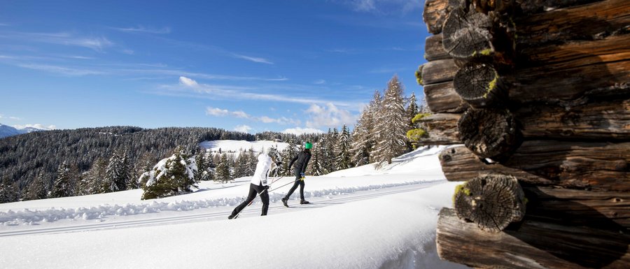 Benvenuti nell’Area Vacanze Sci e Malghe Rio Pusteria! Due sciatori di fondo su pista innevata accanto a una baita di legno