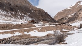 Malga Fane, il più bel villaggio alpino dell’Alto Adige L'immagine mostra un paesaggio montano invernale con un piccolo villaggio circondato da pendii innevati e in parte senza neve. In primo piano scorre un ruscello e sullo sfondo si possono vedere alcune baite di legno e una piccola chiesa.