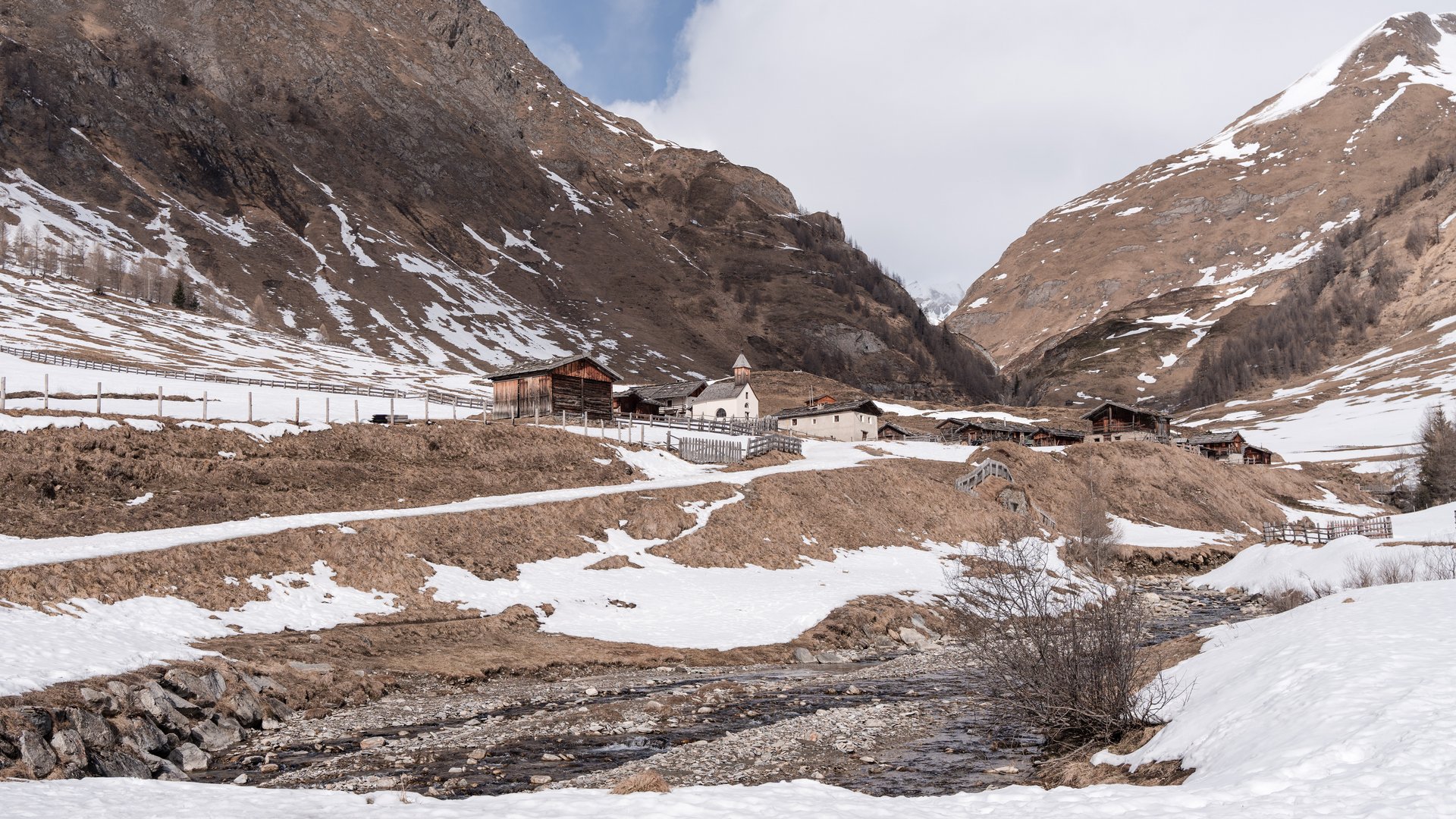 Malga Fane, il più bel villaggio alpino dell’Alto Adige L'immagine mostra un paesaggio montano invernale con un piccolo villaggio circondato da pendii innevati e in parte senza neve. In primo piano scorre un ruscello e sullo sfondo si possono vedere alcune baite di legno e una piccola chiesa.