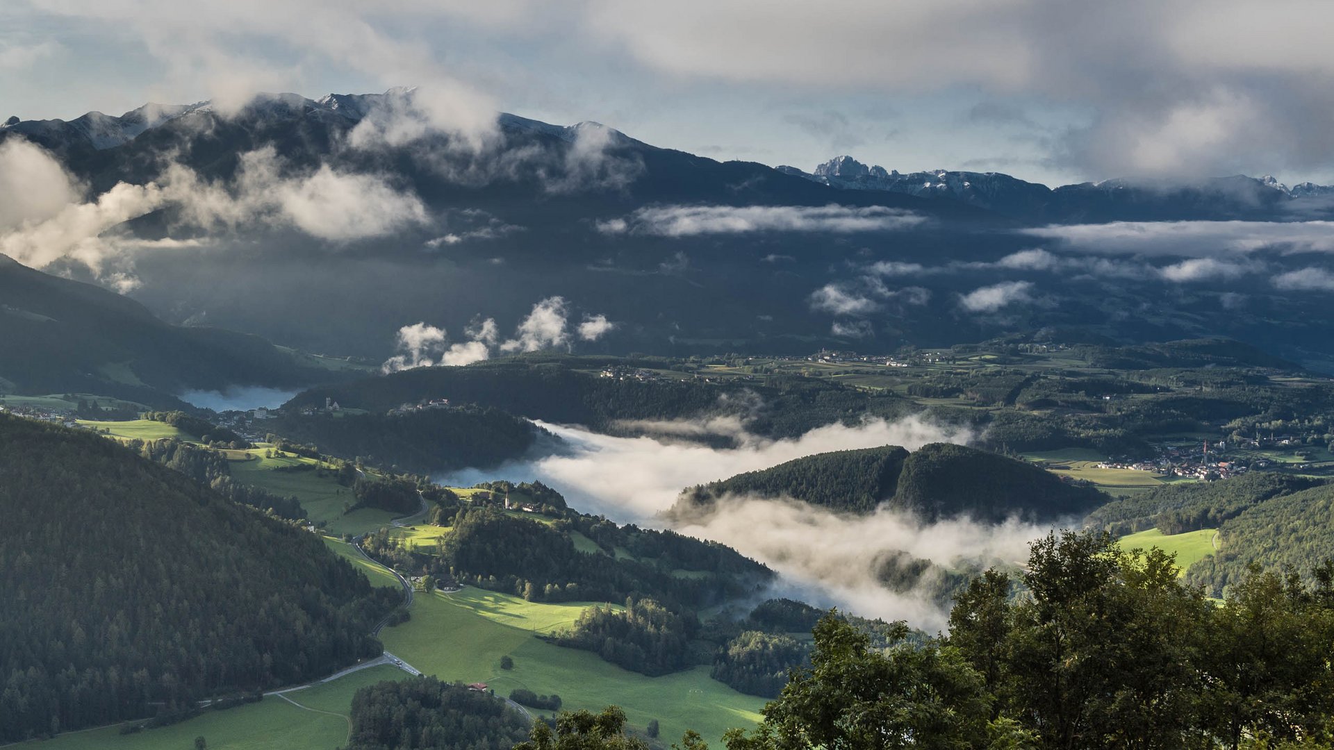 Il paradiso escursionistico di Rodengo L'immagine mostra un vasto paesaggio verdeggiante con dolci colline e valli, attraversate da nuvole nebbiose. Sullo sfondo si ergono montagne scure verso il cielo, parzialmente coperte dalle nuvole, mentre il sole basso illumina la scena.