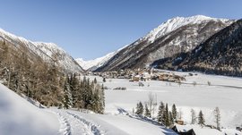 Valles & il villaggio alpino più bello dell’Alto Adige L'immagine mostra un paesaggio invernale coperto di neve con un piccolo villaggio nella valle, circondato da alte montagne. Un sentiero stretto con impronte di piedi attraversa la neve, mentre sullo sfondo si vede un cielo limpido e azzurro.