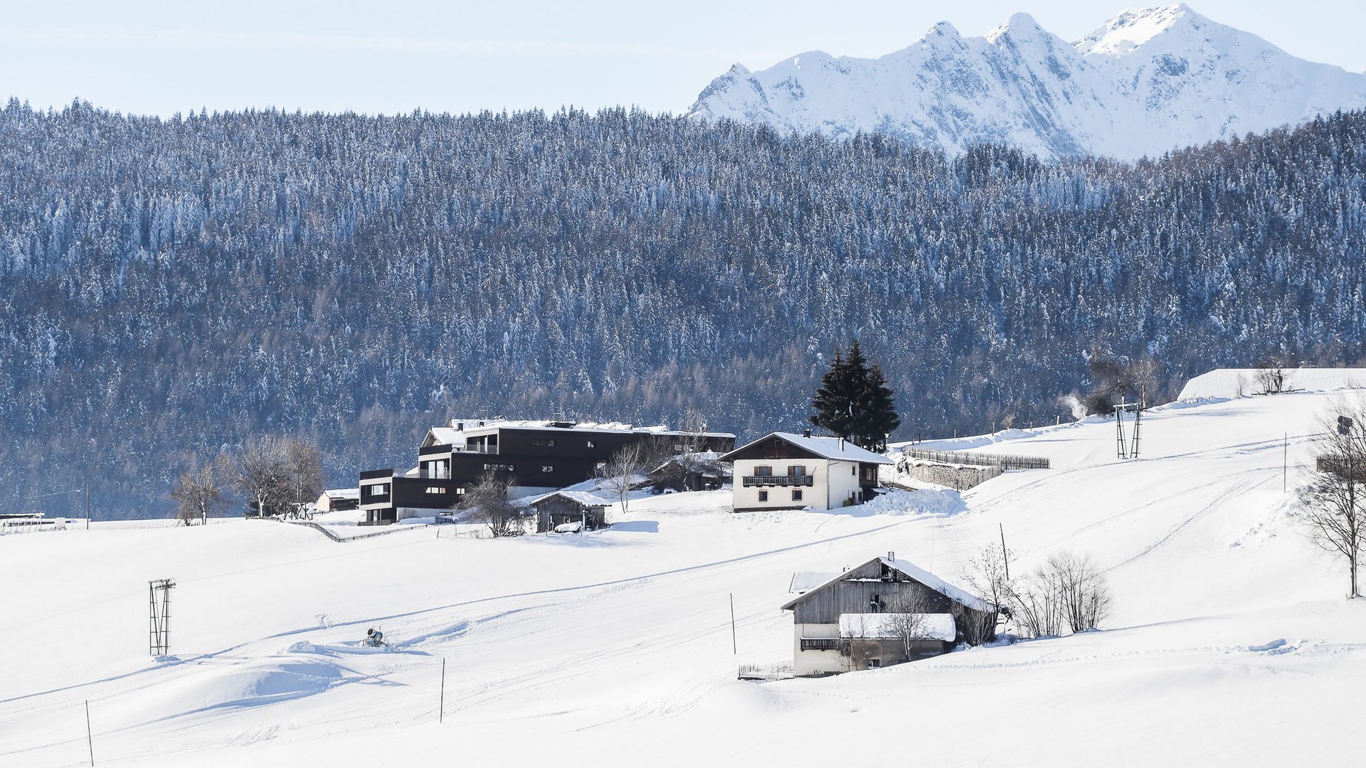 Conoscete già Maranza? L'immagine mostra un paesaggio invernale con colline innevate e edifici, circondati da una fitta foresta innevata e alte montagne innevate sullo sfondo. Le case sono sparse sulle colline e sono in parte moderne, in parte tradizionali.